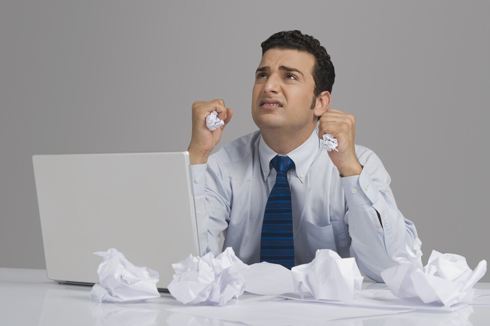 Man suffering while writing; sitting in front of laptop with crumpled paper and clinched fists